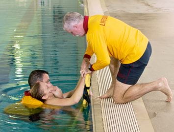 image of lifeguard with casualty in the water while another lifeguard kneels next to the pool placing a spineboard in the water