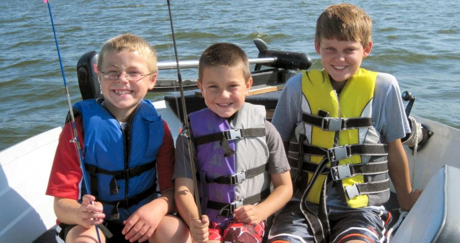 Three young boys wearing lifejackets, in a boat, holding fishing rods