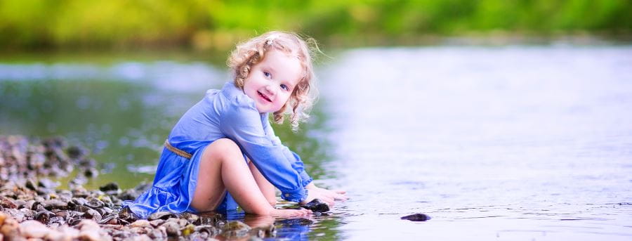 A toddler girl in a lilac dress sitting by the river playing in the water